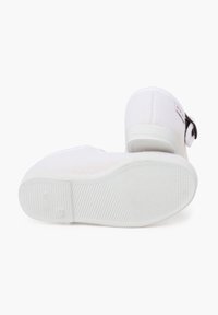 Zapato slip-on de textil blanco con parte superior de punto texturizado, punta redondeada y suela de goma con un sutil patrón de agarre. Detalle de cierre con botón ajustable.
