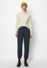 Krēmkrāsas ribiņots turtlenecks džemperis, tumši zili velkamo bikses un melnas masīvas kurpes. Spilgtas dzeltenas berete ar etiķeti.