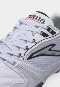 Λευκό ποδοσφαιρικό παπούτσι Joma Dribling με μαύρο λογότυπο, υφασμάτινο συνθετικό επάνω μέρος με υφή και λευκά κορδόνια.