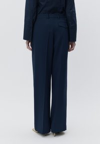 Calças largas azul-marinho com frente lisa, passantes para cinto e bolso traseiro. Tecido com textura lisa. Usadas com saltos bege.