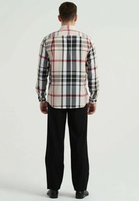 Chemise à manches longues pour homme dans un motif à carreaux beige, noir et rouge, avec des poignets à bouton et un col classique, associée à un pantalon noir.