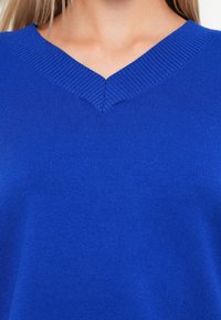 Plava v-neck pulover s rebrastim ovratnikom, izrađen od mekog pletenog materijala. Ima glatku teksturu i ravnomjernu boju.