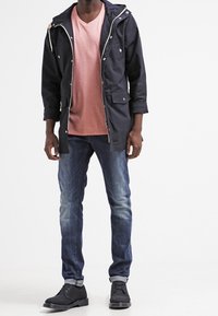 Granatowa parka z kapturem, różowa koszulka z dekoltem w serek, dżinsy i czarne buty. Charakteryzuje się zamkami, kieszeniami oraz lekkim materiałem.