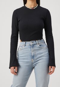 Kvinna som bär en svart ribbad långärmad tröja, ljusblå högmidjade jeans och ett blommigt pärlhalsband med ett rött pärlmitten.