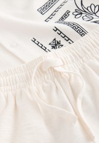 Primer plano de la cintura elástica blanca con cordón atado sobre tela texturizada, junto a tela blanca con patrones geométricos y florales negros.