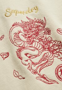Kremowa tkanina z wyraźnym haftem czerwonego smoka z detalami łusek i akcentami kwiatowymi. Nazwa marki "Superdry" jest wyszyta złotym kolorem.