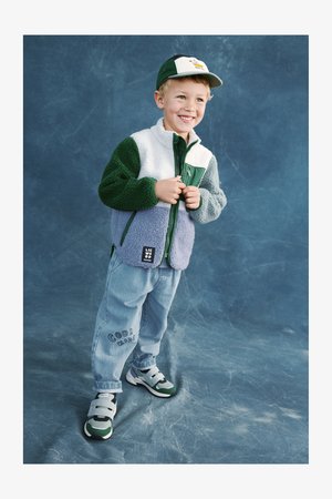 Gyermek, aki több színű (zöld, fehér és levendulalila) polárdzsekit visel, világoskék farmernadrággal és zöld, tépőzáras sportcipővel.