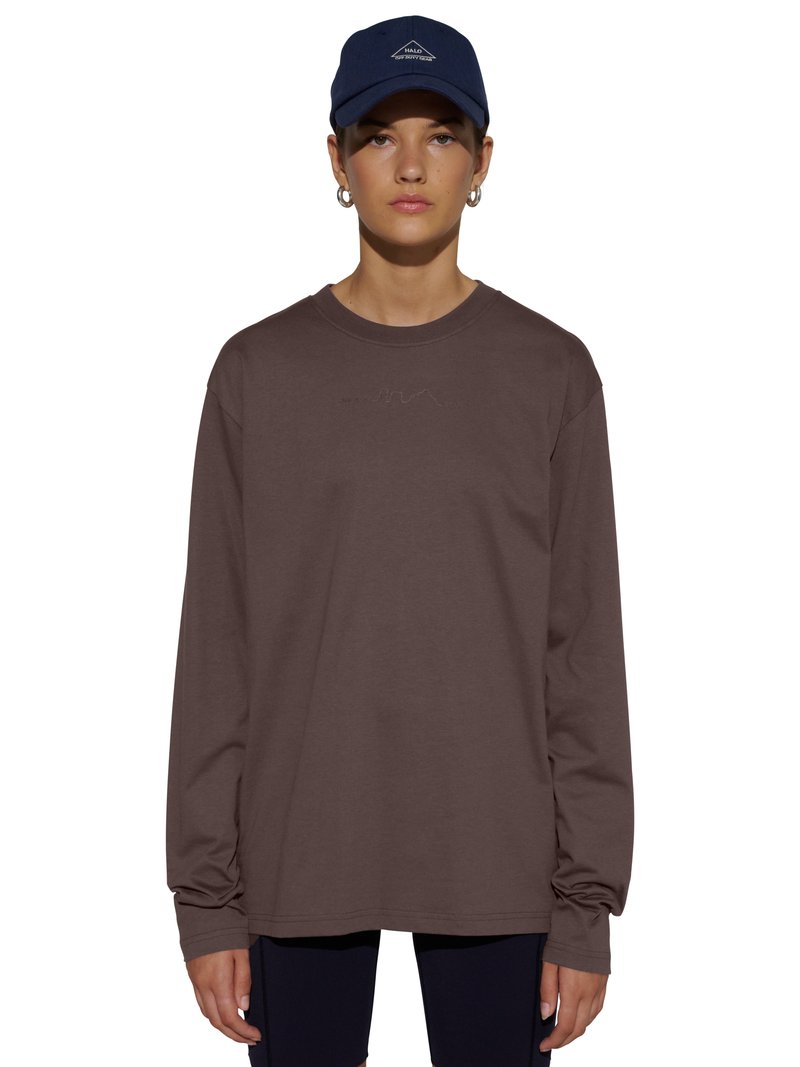 Jeune personne portant un t-shirt à manches longues marron foncé et une casquette bleu marine avec le texte « HALO SOFT WORKWEAR », debout sur un fond blanc.