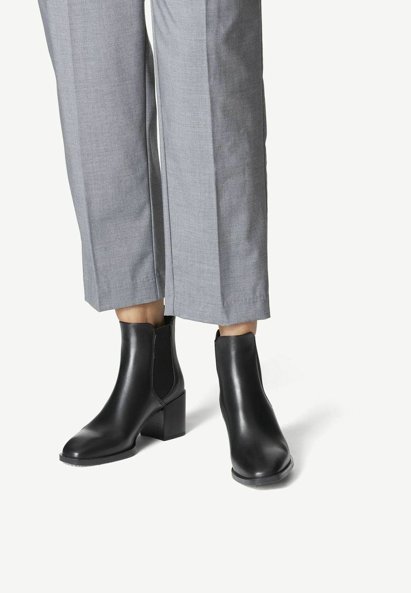 Jambes portant un pantalon gris associé à des bottines en cuir noir avec talons blocs et panneaux latéraux élastiques.