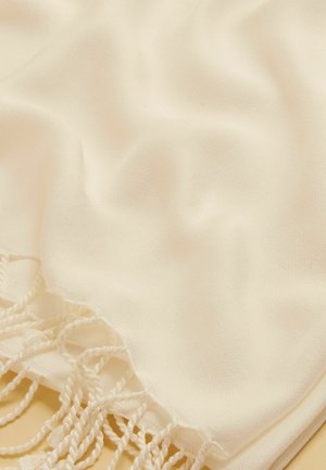 Kreemikas kangas, pehme tekstuuriga ja ühe serva ääres keerutatud sõlmedega, suures plaanis õrnade voltidega.