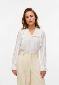 Witte lange mouwen blouse met ruffle accenten en kantdetails bij de halslijn, gecombineerd met hoge crèmekleurige trousers met knopen.