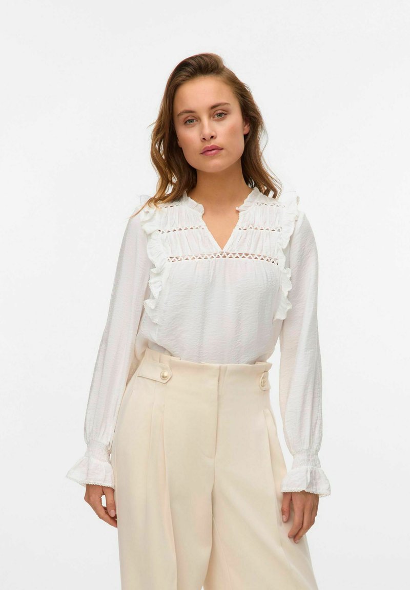 Witte lange mouwen blouse met ruffle accenten en kantdetails bij de halslijn, gecombineerd met hoge crèmekleurige trousers met knopen.