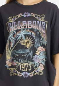 Μαύρη μπλούζα με μια πολύχρωμη γραφική απεικόνιση που περιλαμβάνει το "BILLABONG", τον ήλιο, τροπικά φυτά και λουλούδια, καθώς και το κείμενο "ΑΝΑΣΤΗΣΟΥ ΜΕ ΤΟΝ ΗΛΙΟ 1973."
