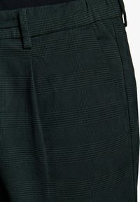 Grüne karierte Hose aus strukturiertem Stoff, mit einem Bund und Taschen an den Seiten. Das Muster besteht aus kleinen Quadraten und horizontalen Linien.