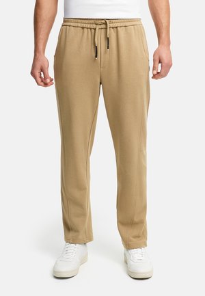 Homme portant un pantalon de survêtement beige à cordon et des baskets blanches, debout les bras détendus le long du corps sur fond blanc.