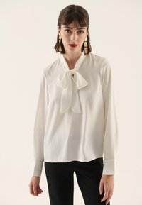 White blouse with a tie neckline, long sleeves, and smooth texture, paired with black trousers. Minimalist design, no patterns or accents.