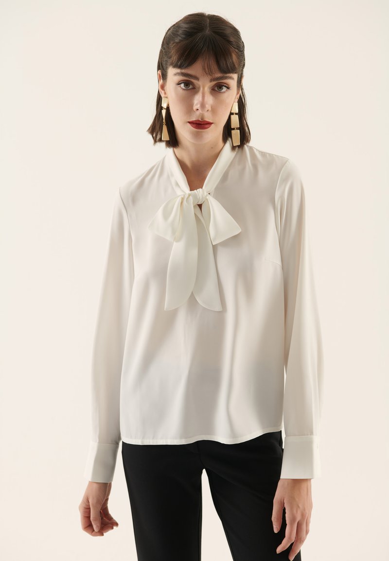 White blouse with a tie neckline, long sleeves, and smooth texture, paired with black trousers. Minimalist design, no patterns or accents.