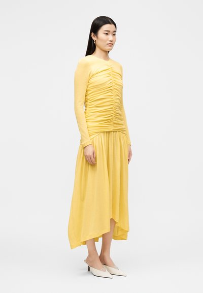 Femme portant un haut jaune à manches longues froncé et une jupe midi asymétrique assortie avec des mules blanches à talons, debout devant un fond uni.