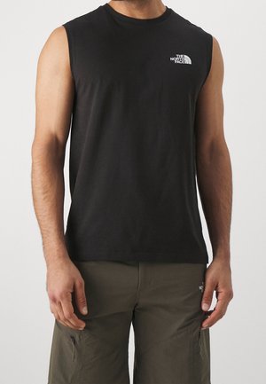 Homme portant un débardeur noir The North Face et un pantalon cargo vert olive, debout devant un fond clair uni.
