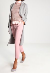 Světle šedý dlouhý kabát, růžové přizpůsobené kalhoty a texturovaný růžový svetr s volánkovým detailem. Doplňují to světle šedé boty na vysokém podpatku.