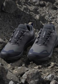 Ζευγάρι μαύρα και γκρι παπούτσια trail running Adidas Terrex τοποθετημένα σε τραχιά, βραχώδη επιφάνεια στο ύπαιθρο.