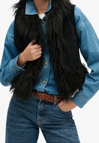 Gilet in pelliccia sintetica nera sopra una camicia di jeans, con un design testurizzato e scostante. Indossato con una cintura marrone intrecciata e jeans blu.