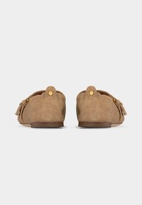 Paire de mocassins beige en daim avec détail de bouton doré sur les languettes de talon, semelles plates et pampilles latérales, vus de dos sur fond blanc.