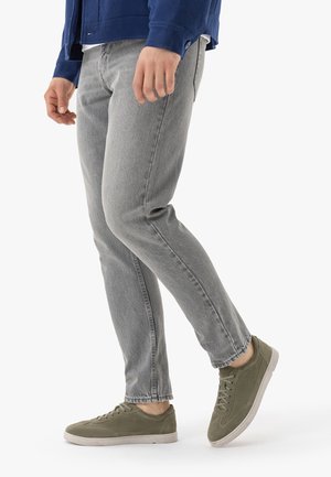 Persona che indossa jeans grigi e scarpe da ginnastica stringate verde oliva, in piedi con un piede leggermente sollevato.