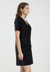 Robe noire avec des manches courtes, un décolleté en V et un design à ourlet superposé. Fabriquée dans un tissu lisse avec une silhouette ajustée.