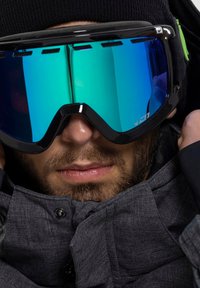 Black ski goggles with reflective blue and green lenses, worn by a person with a short beard, in a gray jacket with a high collar.