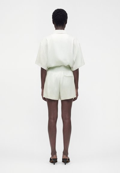 Personne debout, dos tourné, portant une chemise blanche ample à manches courtes, un short blanc taille haute et des sandales à talons noires sur fond uni.