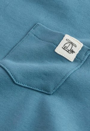 Tessuto color teal con una piccola tasca sul petto dotata di un'etichetta bianca che mostra un design di logo nero. La texture sembra lavorata a maglia e morbida.