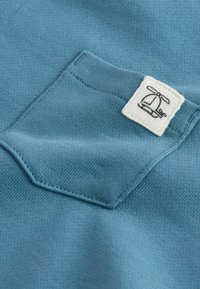 Tissu tricoté bleu avec une petite étiquette blanche carrée montrant une icône noire d'hélicoptère cousue près du bord d'une poche.