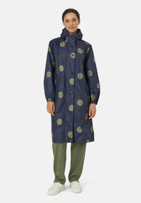 Marineblå regnfrakke med store olivengrønne prikker, med hætte, elastiske cuffs, tryklås og sidelommer. Modellen har den på med grønne bukser.
