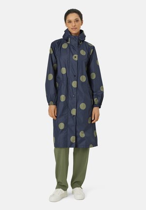 Marineblauer Regenmantel mit großen olivgrünen Polka-Dots, mit Kapuze, elastischen Bündchen, Druckknopfverschluss und Seitentaschen. Das Modell trägt ihn zu grünen Hosen.