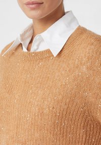 Frau in einem beigen gestrickten Pullover mit kleinen Pailletten über einem weißen Kragenhemd, das den Ausschnitt und das untere Gesicht zeigt.