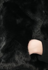 Čierna umelá kožušina s jemnou, hustou textúrou. Ruka s rebrovaným čiernym rukávom je čiastočne viditeľná proti kožušine.