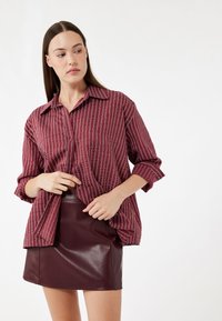 Burgundowa, pasiasta koszula zapinana na guziki z kołnierzykiem, z podwiniętymi rękawami, połączona z dopasowaną burgundową mini spódnicą z ekoskóry. Gładka tekstura.