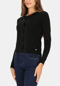 Czarny dzianinowy kardigan z okrągłym dekoltem, zapięcie na guziki z przodu oraz falbaniastym wykończeniem na środku. Połączony z ciemnymi dżinsami.
