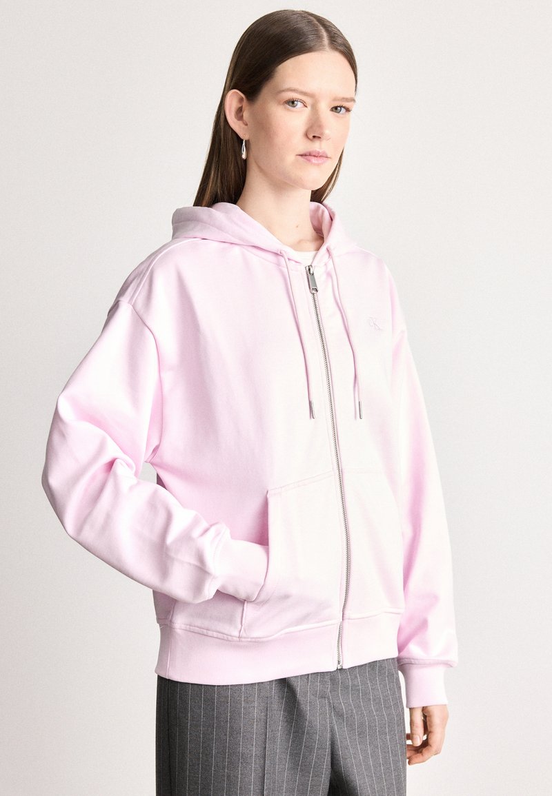 Svetlo ružová mikina na zips vyrobená z mäkkého materiálu s veľkou prednou kapsou a kapucňou na šnúrku; obsahuje jednoduché vyšívané logo.