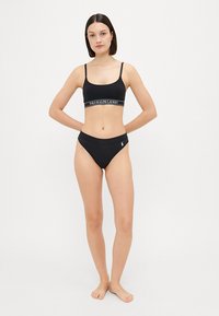 Black bikini set made of smooth fabric. Bralette features logo band; bottom has a low rise with a side logo accent. Simple, fitted design.