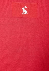 Tejido de punto rojo con un pequeño logotipo de conejo bordado en blanco sobre un parche rectangular. La textura es suave y uniforme.
