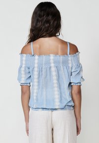 Top azzurro chiaro a spalla scoperta con bordi arricciati, tessuto texture e ricami verticali bianchi. Spalline sottili e vestibilità ampia.