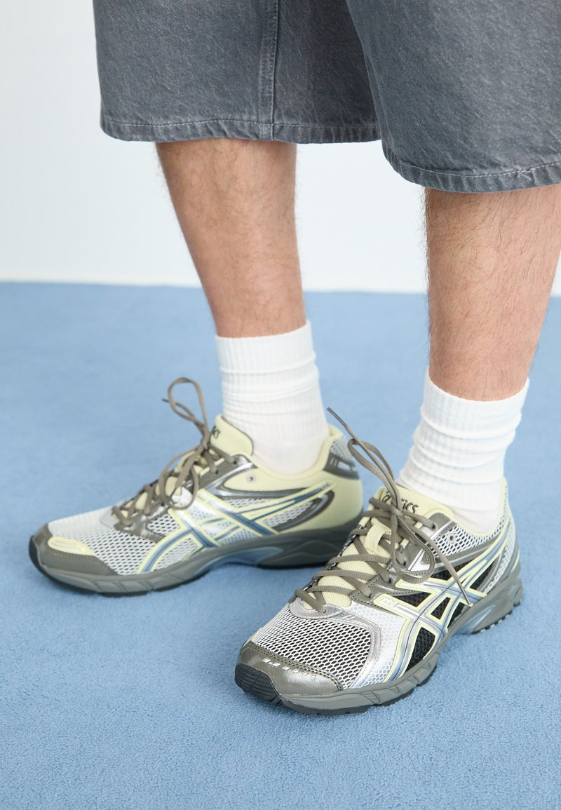 Lichtgrijze sportschoenen met een bovenkant van mesh, synthetische accenten, reflecterende details en een grijze zool, in combinatie met witte crew-sokken.