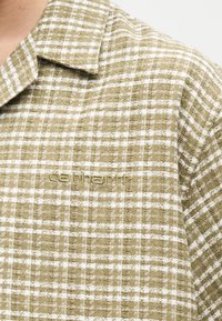 Κοντινή εικόνα πουκάμισου από ύφασμα καρό σε ανοιχτό καφέ και άσπρο, με γιακά και κεντημένο το λογότυπο Carhartt στο στήθος.