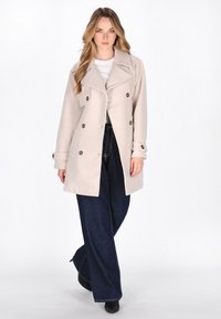 Beige dubbelzijdig trenchcoat met grote knopen en brede revers, gecombineerd met donkerblauwe wijde jeans en zwarte enkelboots.