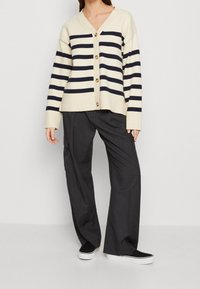 Gestreepte cardigan in crème en marineblauw met een V-hals, houten knopen, geribbelde manchetten, gecombineerd met losse grijze broek en zwarte sneakers.