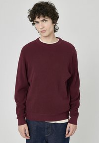 Pull en tricot côtelé bordeaux avec un col rond et des manches longues, associé à une chemise de couleur claire en dessous et un jean foncé.