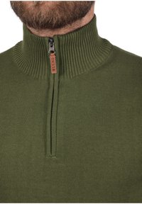 Grüner Zip-Hoodie mit hohem gerippten Kragen, der einen kontrastierenden Reißverschluss-Zipper und einen strukturierten Strickstoff aufweist.