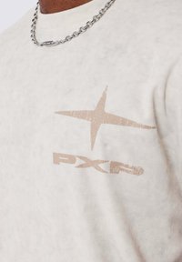 T-shirt in cotone color crema con finitura testurizzata, caratterizzato da una grafica beige sbiadita di una stella e dal logo "PXP" sul petto. Catena argentata visibile.